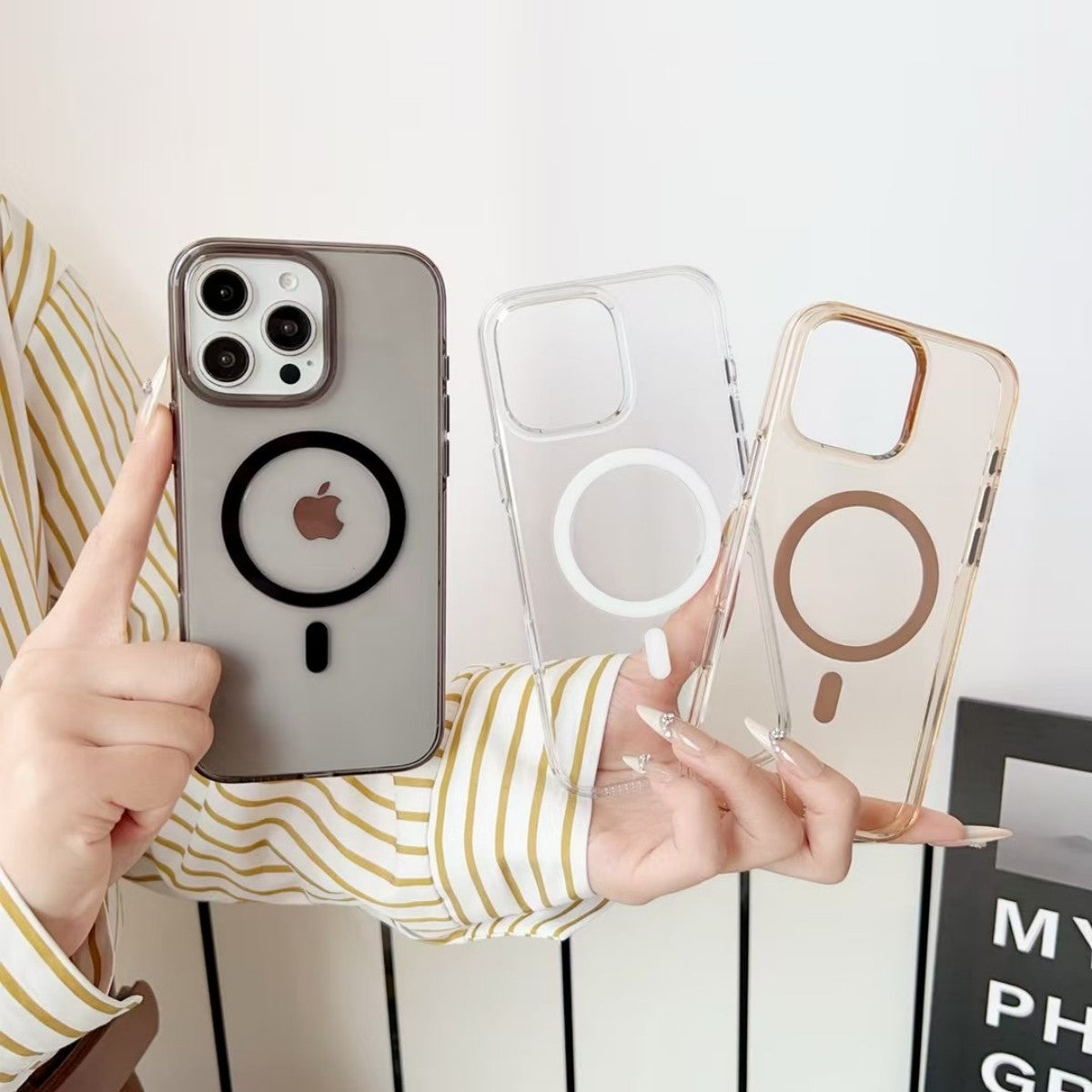 Three AuraGuard Series iPhone Clear Cases in black, white, and gold MagSafe ring variants, displayed in hand for color comparison.