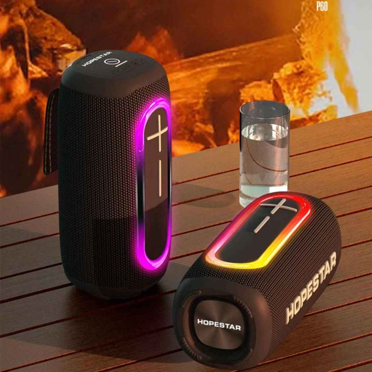 Hopestar P60 Outdoor Bluetooth Speaker with RGB lighting, placed on a wooden table near a fire for an outdoor ambiance.